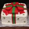 CH-022 is a square cake designed to look like a gift, with white icing, green and red fondant ribbons and bow, plus holly decorations on the sides. Presented on silver foil atop a wooden surface.