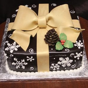 CH-021 is a square cake styled as a gift, featuring dark chocolate fondant, a large gold fondant bow, white snowflakes, fondant pinecone, holly leaves and berries, displayed on a silver-patterned board.