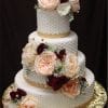 The WD-078 is a three-tiered white cake with textured icing, adorned with large pink, burgundy, and white flowers, gold accents, and greenery. It sits on a gold-patterned base against a dark background.