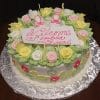 BB-001 is a round cake topped with pink, yellow, and white icing roses, green leaves, baby shoe toppers, and a “Welcome Sophia Luci” sign in pink script. Displayed on a silver-patterned board.