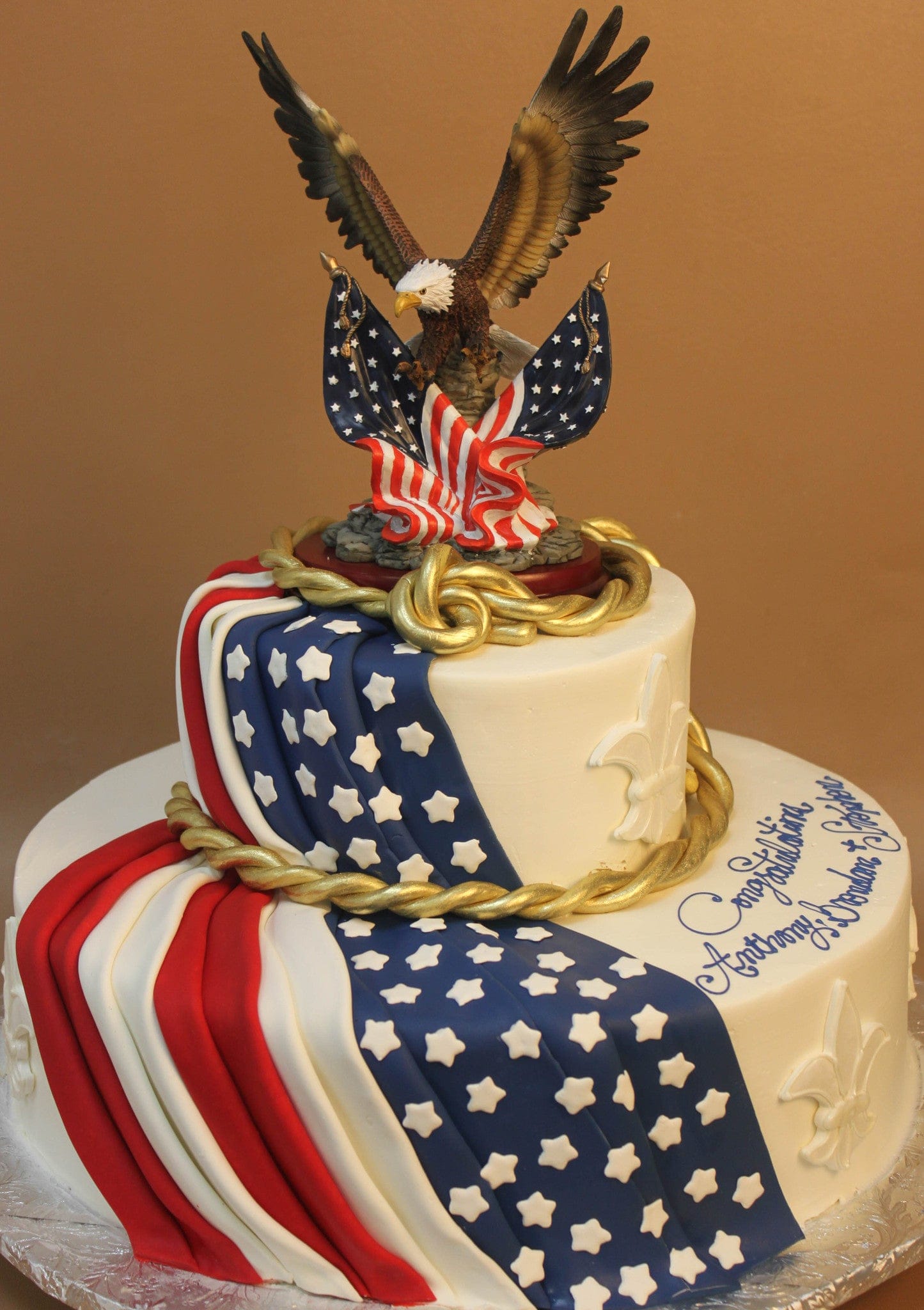 The TC-152 is a two-tiered cake featuring an American flag design, gold rope accents, white stars, and a bald eagle figurine with spread wings on top. A congratulatory message decorates the side.
