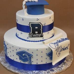 TC-131 is a two-tiered white graduation cake with blue ribbons, blue cap decorations and topper, a scroll with “Congratulations Michael,” plus a large “B” and “Wamps” on the front.
