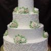 The WC-064 is a four-tiered white wedding cake featuring intricate swirls, dotted accents, and white rose flowers with green leaves on each tier. It sits on a gold base against a black background.