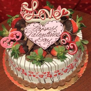 The VC-006 Display Strawberry Grand Marnier cake is a round Valentine’s Day cake topped with chocolate-dipped strawberries, pink and red heart decorations, and a large heart-shaped plaque that reads "Happy Valentines Day" with "Love" on top.