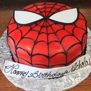 A CB-006B round birthday cake decorated as Spider-Man’s face features red icing, a black web pattern, large white eyes, and “Happy Birthday Nicholas” written in cursive on a fondant strip.