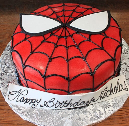 A CB-006B round birthday cake decorated as Spider-Man’s face features red icing, a black web pattern, large white eyes, and “Happy Birthday Nicholas” written in cursive on a fondant strip.