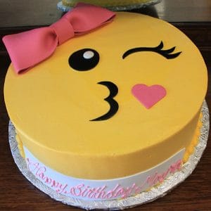 The CB-005A is a round yellow cake designed as a winking emoji with a pink bow on top and a pink heart on the cheek. "Happy Birthday" is written in pink icing on the white cake base.