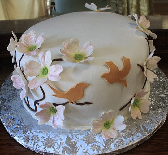 The BR-040 is a round cake with white fondant, adorned with pink and white flowers and tan bird-shaped cutouts, presented on a silver floral-patterned cake board.