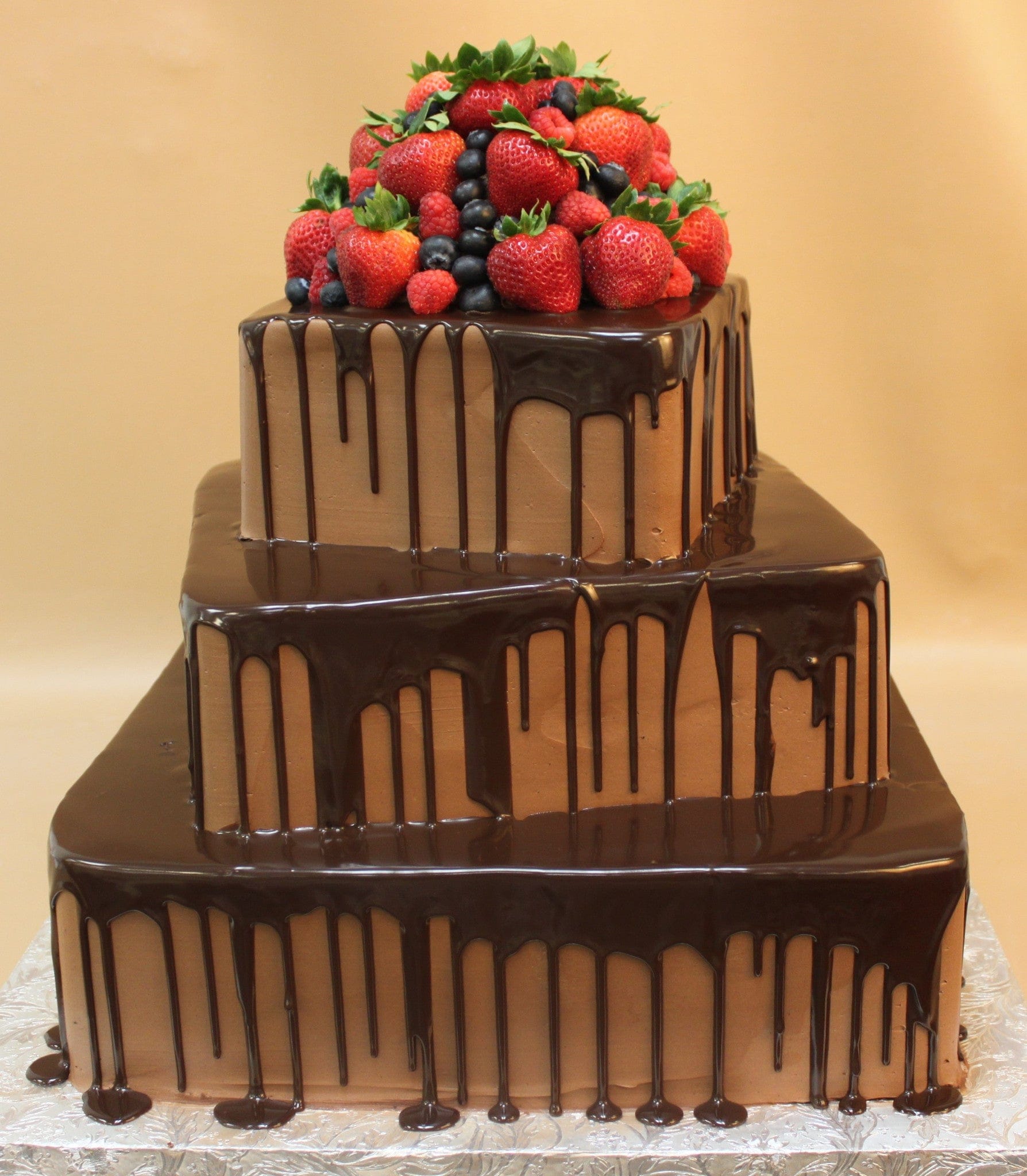 The TC-140 is a three-tier square chocolate cake with chocolate ganache drips, topped with fresh strawberries, blueberries, and raspberries, set against a beige background.