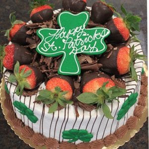The STP-004 cake features chocolate and cream frosting, chocolate-dipped strawberries, chocolate shavings, green shamrocks, and a large green shamrock topper with “Happy St. Patrick’s Day.”.
