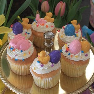 Five EA-041 Display Easter Decor Gold cupcakes with chocolate filling, topped with pink and purple eggs, yellow chicks, and bunnies, are arranged on a gold stand surrounded by fresh tulips and daffodils.