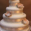 WD-209 is a four-tiered white cake adorned with gold ribbons, silver bead accents, and large jeweled gold flower toppers, displayed against a brown background.