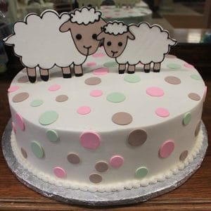 BB-046 is a round cake adorned with pink, green, and brown polka dots, featuring two cartoon sheep figures standing side by side on top. The cake sits on a silver board.