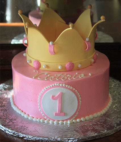 The CB-016D cake features pink icing, a large yellow crown topper, white and pink pearls, a white circle with the number 1 on the front, and "Victoria" written in white icing.