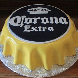 The BD-025 Corona cake is shaped like a yellow bottle cap with the Corona Extra name and crown logo on black, mimicking the beer's branding. It’s presented on a patterned silver board.