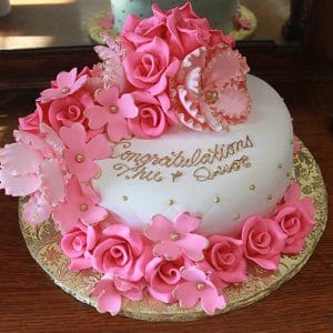 The BD-054C pink quilted is a round white cake adorned with pink fondant flowers and gold pearls. "Congratulations Thu & Quoc" is written in gold icing on top, and the cake sits on a gold-patterned board.