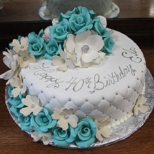 The BD-054A Aqua quilted is a round white cake with quilted icing, adorned with teal and white fondant flowers. "Happy 40th Birthday Sue" is written in silver icing on top, and the cake rests on a silver cake board.