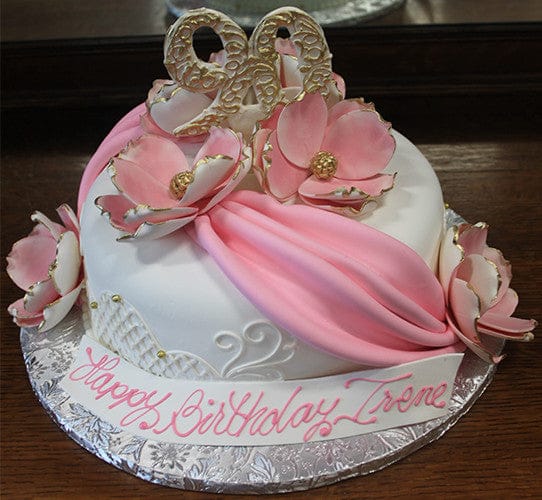 The BD-055B Pink Swag is a round white birthday cake with pink draped fondant, gold-edged pink flowers, a gold "90" topper, and "Happy Birthday Irene" written on the base.