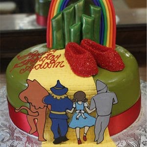 The BD-079 cake is Wizard of Oz-themed, with Dorothy, Scarecrow, Tin Man, and Lion on a yellow brick road toward ruby shoes, green towers, and a rainbow. "Happy Birthday" is written in red icing.