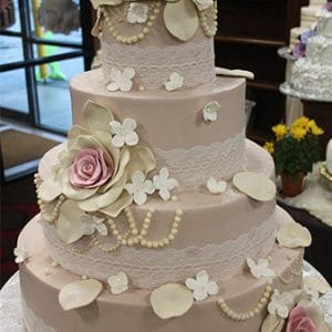 The WD-066 is a three-tier blush pink wedding cake adorned with large pink and white sugar roses, lace icing, white sugar pearls, and small white flower accents, beautifully displayed indoors with floral arrangements nearby.
