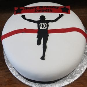 The ST-012 Running cake is round and white, featuring a silhouette of a runner with a "40" bib crossing a red finish line. A red banner on top says "Happy Birthday Durgesh" with black stars at each end.