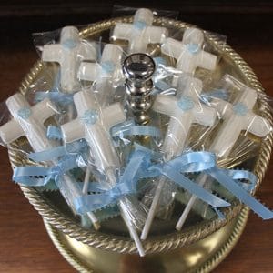 RC-115: White chocolate cross lollipops with blue ribbon bows and blue flower accents, individually wrapped in clear plastic and displayed in a gold bowl with a decorative silver handle.