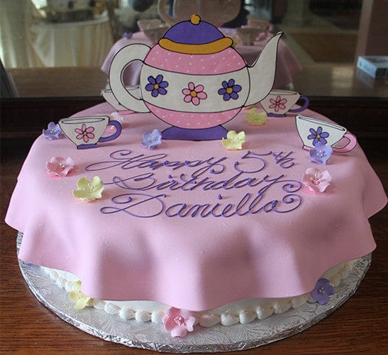 The CB-028D is a round cake with pink fondant, "Happy 5th Birthday Daniella" in purple icing, plus paper teapot and teacups, and pastel fondant flowers as decorations.
