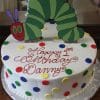 The CB-009X is a round birthday cake adorned with colorful polka dots, a Very Hungry Caterpillar topper, and "Happy 1st Birthday Danny" written in red icing on a foil-covered board.
