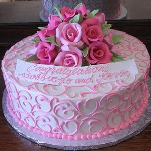 The BR-010 Shades of Pink Roses is a round cake with pink swirls, a pink icing border, and a bouquet of pink and purple roses on top. A white banner says, “Congratulations Bobbie Jo and Kevin.”.