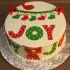 The CH-017 is a festive Christmas cake with JOY in red and green icing, yellow dots, a garland of stockings, red bows, and a red bird on top, presented on a silver foil-covered board.
