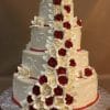 WC-050: A four-tiered white wedding cake with intricate white icing, adorned with cascading red and white roses and flower accents, displayed on a silver board against a brown background.
