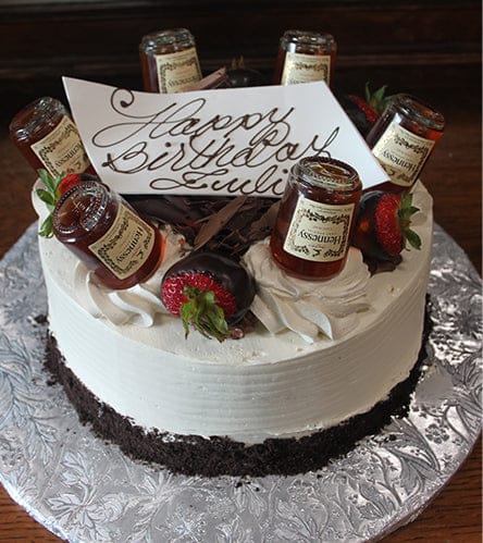 The BD-024H Hennessy birthday cake is round with white frosting, chocolate shavings, chocolate-dipped strawberries, mini Hennessy bottles on top, and a "Happy Birthday Julius" card in cursive, presented on a silver board.