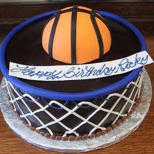 The ST-005 Basketball cake is a round chocolate cake with white icing in a basketball net pattern, topped with a fondant basketball and a white "Happy Birthday Ricky" banner in blue icing.