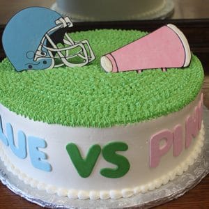 The BB-051 cake is round with green icing, topped with a blue football helmet and a pink megaphone. "BLUE VS PINK" is written in colorful letters on the side.