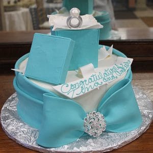 BR-018: A blue and white cake designed as a gift box, topped with a fondant bow, jeweled brooch, and edible ring. Features "Congratulations Zina and Amber" written in blue icing.