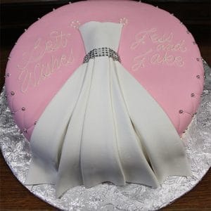 BR-039 is a round cake with pink icing, a white fondant dress centerpiece, silver beads, and "Best Wishes Jess and Jake" in yellow icing, displayed on a silver floral-patterned board.