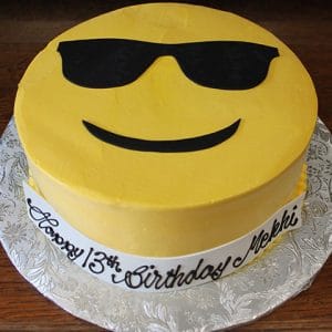 The CB-005D cake is a round yellow cake featuring a cool sunglasses emoji face, displayed on a silver platter, with "Happy 13th Birthday Mekhi" written in black icing on a white strip around the side.