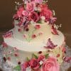 TC-092: A two-tier white cake adorned with pink and red fondant roses, green leaves, small white flowers, pink butterflies, and red ladybugs on a patterned silver board against a brown background.
