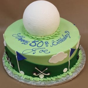 A round ST-007B Golf Ball birthday cake topped with a large white golf ball, green icing, blue and white flags, clouds, golf clubs, and "Happy 50th Birthday Joe" written in blue icing.