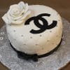 The BD-027C Designer white Rose is a round, quilted white cake with edible silver pearls, a large fondant rose, a black bow and Chanel logo on top, presented on a silver floral-patterned board.