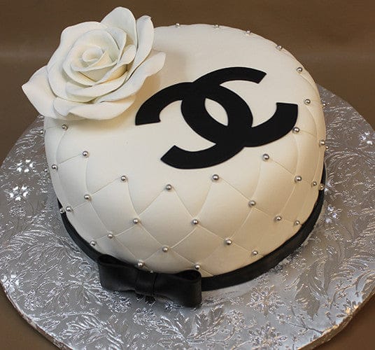 The BD-027C Designer white Rose is a round, quilted white cake with edible silver pearls, a large fondant rose, a black bow and Chanel logo on top, presented on a silver floral-patterned board.