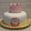 CB-016F is a white cake with a pink crown topper, gold bead trim, "Ava" in pink on a plaque with pink roses and a bow, displayed on a gold cake board against a wooden background.