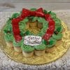 The CC-082 Christmas Wreath features chocolate cupcakes with white filling, decorated festively with green frosting, red holly berries, a big red bow, and a "Merry Christmas" plaque, artfully arranged on a gold tray.