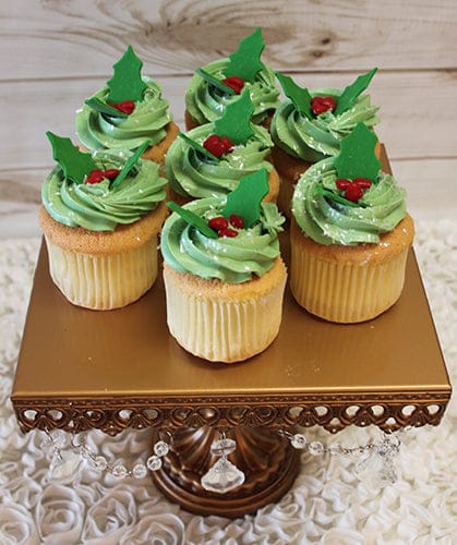 The CC-076 Gold cupcake features chocolate filling and holly berry decor, displayed on a gold ornate stand with crystal accents against a white textured surface and wooden wall background.