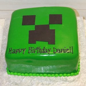 A square green CB-011F cake decorated as a Minecraft Creeper face with "Happy Birthday Daniel!" in black icing on the front, displayed on a white patterned board.