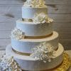 The WC-003 is a three-tiered white wedding cake adorned with gold ribbon, pearl-like dots, and white sugar roses, set on a gold cake board against a wooden background.