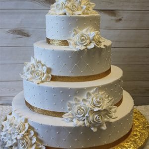 The WC-003 is a three-tiered white wedding cake adorned with gold ribbon, pearl-like dots, and white sugar roses, set on a gold cake board against a wooden background.