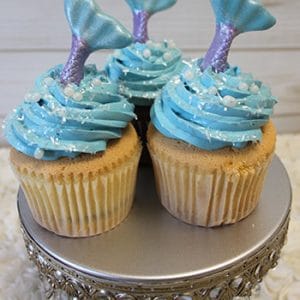 Three CC-026B chocolate cupcakes with white chocolate mousse filling, decorated with white pearls and shimmering mermaid tail toppers, displayed on a silver stand with crystal embellishments.