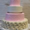 WC-006 is a four-tiered wedding cake with white icing roses on the base, pink and white layers above, pearl-like piping, and a sparkling gold ribbon on the second tier, displayed against a wooden background.