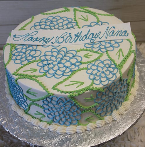 The BD-146 Blue Hydrangea is a round white cake with blue and green floral designs, topped with a white plaque reading "Happy Birthday Nana" in blue cursive, and presented on a foil-covered board.
